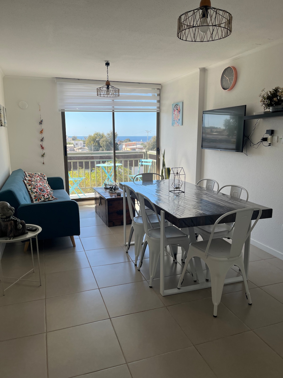 A bright living and dining area features a blue sofa, a wooden dining table with six white chairs, and a mounted television. Large windows allow natural light and showcase a view of the bay. Decorative elements, such as hanging lights and artwork, enhance the space.