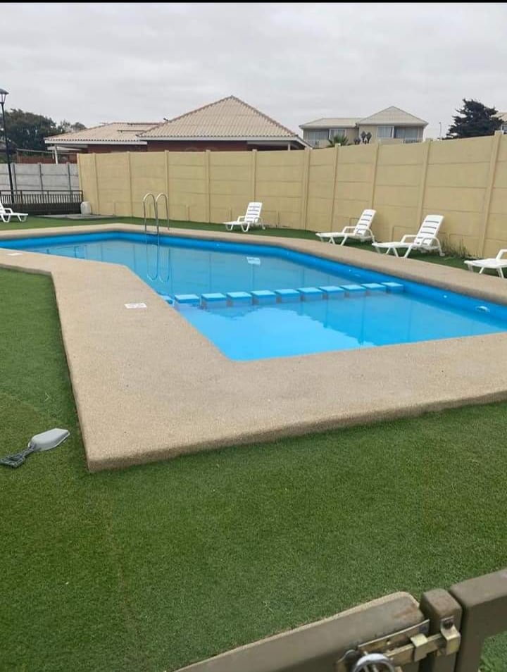 A swimming pool is framed by manicured grass and a surrounding wall. Several lounging chairs are positioned along the pool's edge. The clear blue water reflects a cloudy sky, while the gentle contours of the pool create a welcoming space for relaxation.