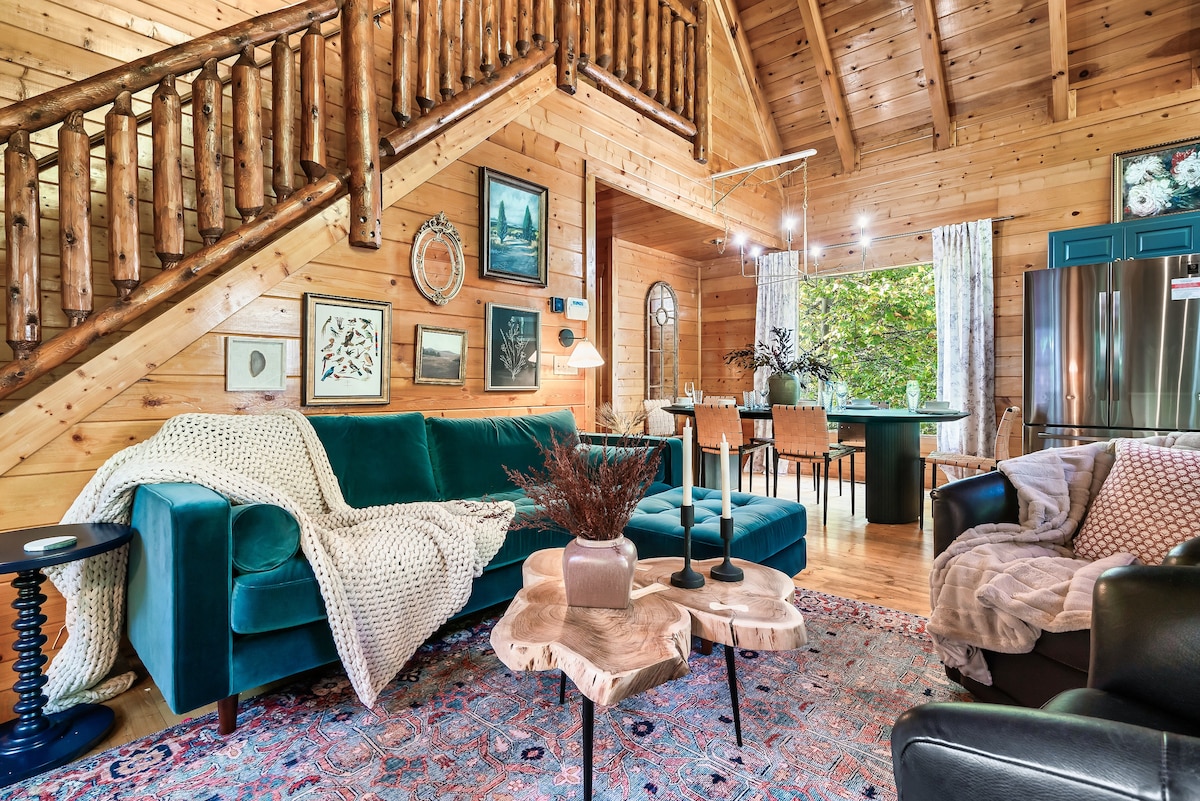 A spacious living area is depicted, featuring a plush teal sofa adorned with a soft blanket and decorative pillows. A natural wood coffee table sits in front, enhancing the inviting ambiance. The walls showcase framed artwork, and large windows allow natural light to illuminate the room.