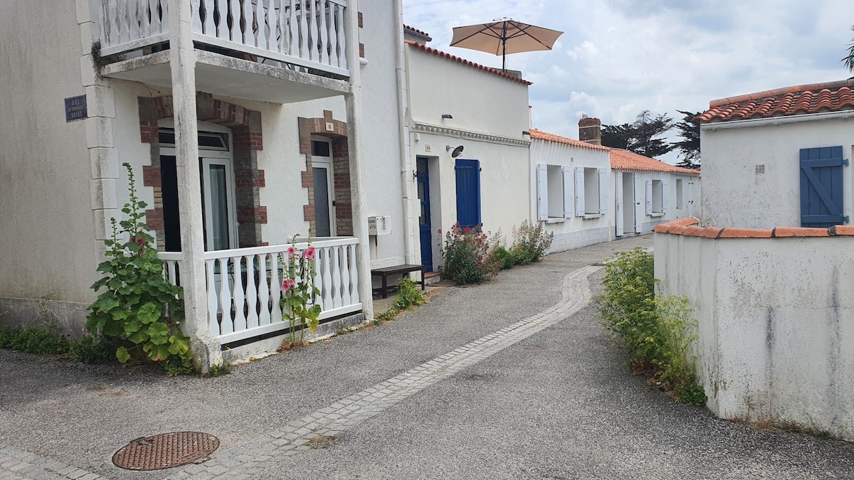 The image depicts a charming pathway winding through a white building complex. Lush greenery and colorful flowers line the sides, complementing the white walls and blue shutters. A patio umbrella shades an outdoor seating area, inviting relaxation on a leisurely day.