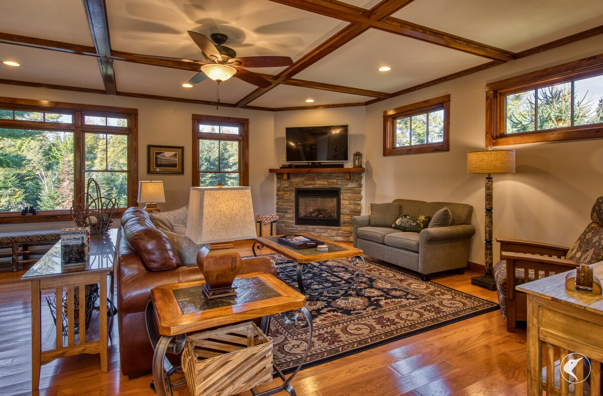 A warm living room is presented with wooden beams and large windows allowing natural light to fill the space. A leather sectional sofa and a comfortable gray couch are positioned around a stone fireplace. A patterned area rug anchors the seating area, enhancing the cozy feel.