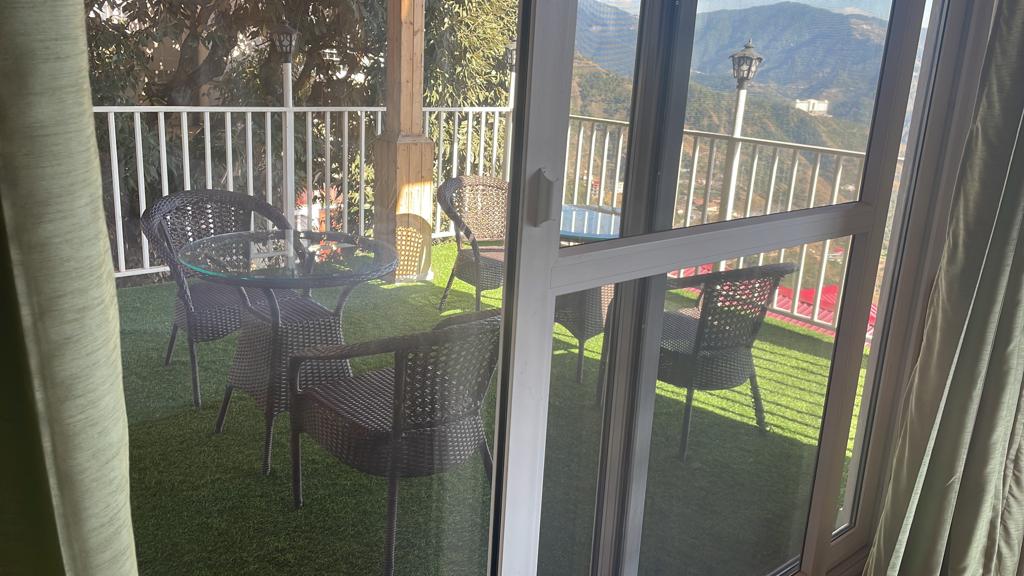 A balcony is featured with artificial grass covering the floor, creating a lush environment. Four wicker chairs surround a glass-top table, providing a comfortable seating arrangement. A scenic view of the mountains is visible through the railing, with soft curtains partially framing the entrance.