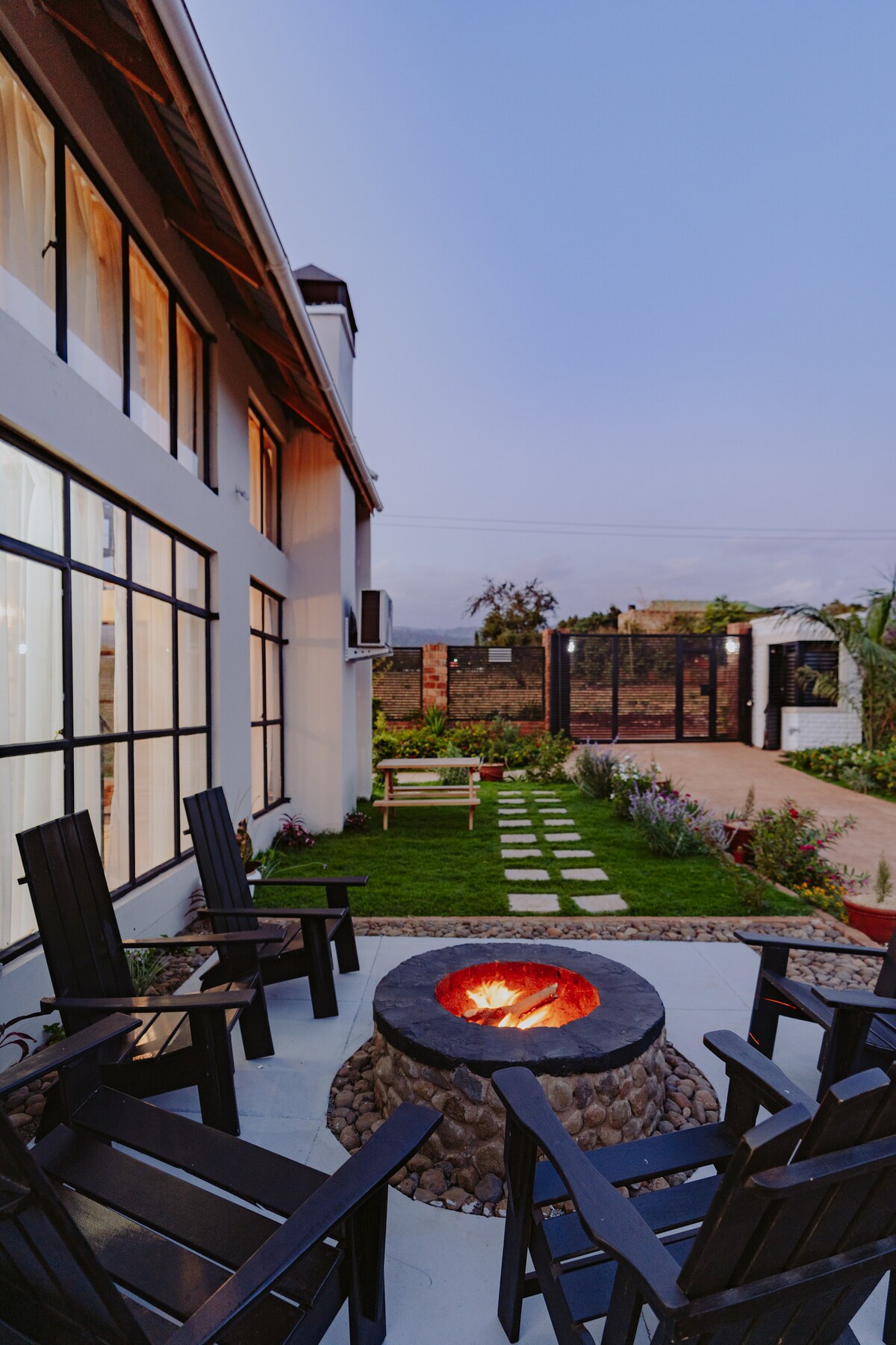 A cozy outdoor seating area surrounds a circular fire pit, set against a backdrop of a modern two-story building. The space features several black wooden chairs arranged invitingly, with a pathway of stones leading towards a beautifully landscaped garden.