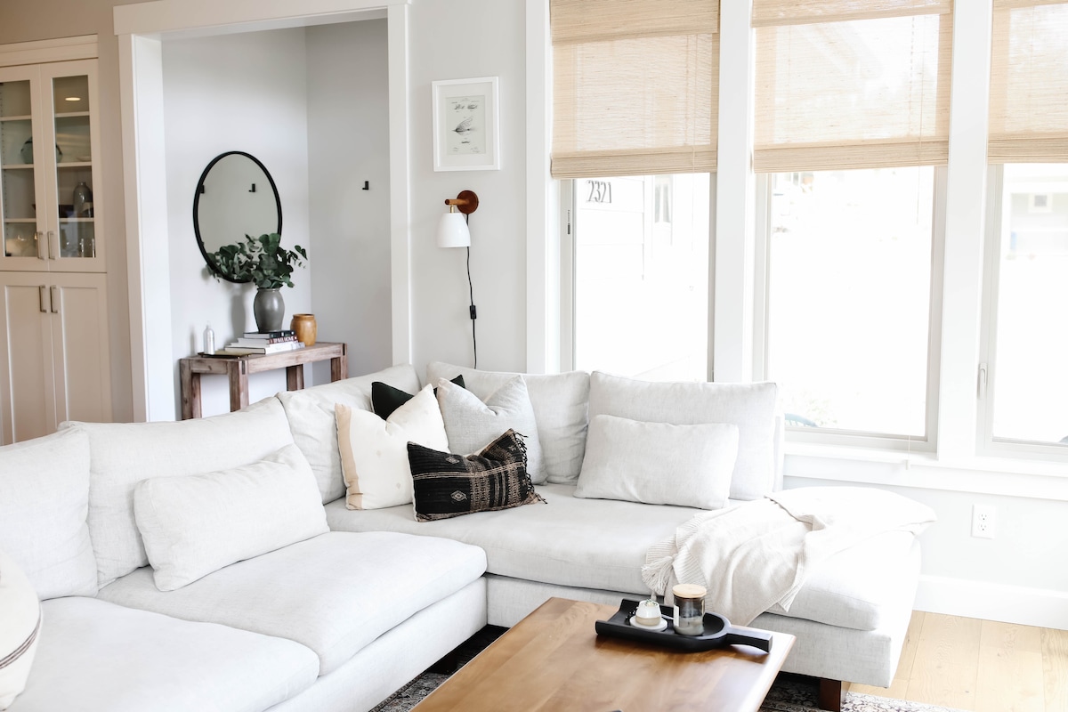 A spacious living area features a light-colored sectional sofa adorned with various pillows. A wooden coffee table sits in front, displaying two cups and a small dish. Natural light filters in through large windows with beige shades, enhancing the clean and modern decor.