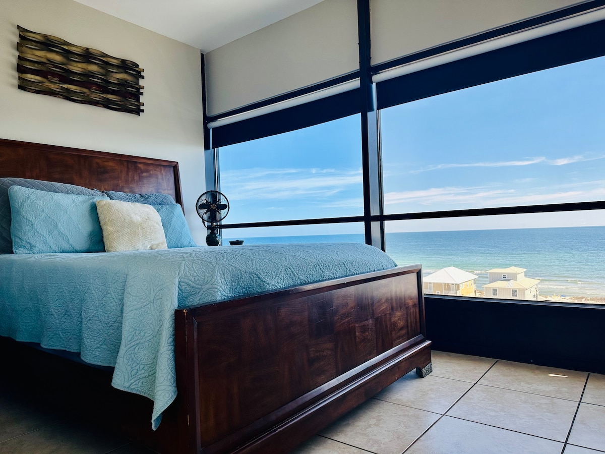 A spacious bed dressed in light blue linens rests adjacent to a large window, offering expansive views of the ocean. Natural light floods the space, enhancing the calming hues of the room. A decorative wall art piece adds texture above the bed.