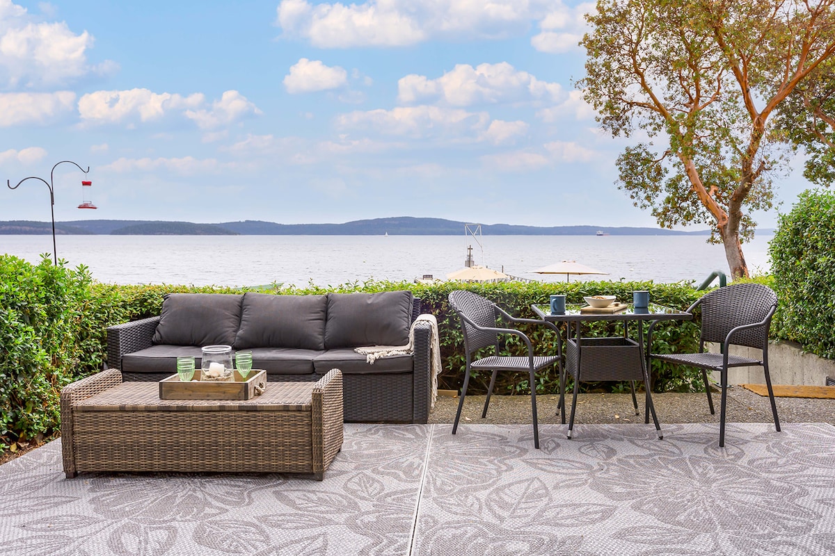 A private patio area features comfortable seating with a dark, cushioned sofa and a wicker coffee table. A dining table with chairs is positioned beside the water, showcasing views of the serene ocean and distant islands, framed by greenery and a partially cloudy sky.