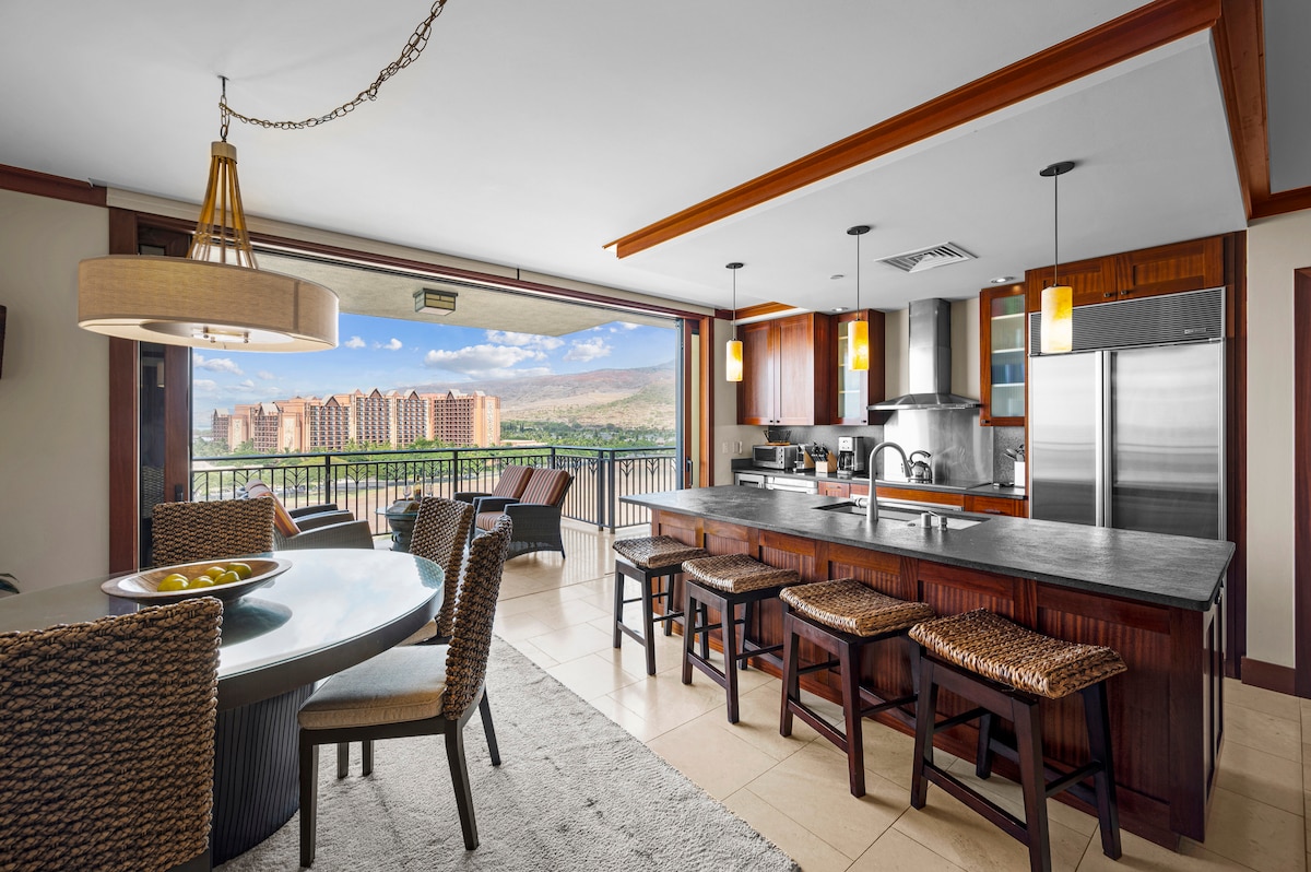 A modern kitchen and dining area are showcased, featuring upgraded stainless steel appliances and a large island with seating for six. Floor-to-ceiling windows provide views of the outdoor lanai and surrounding landscape. The dining table is set for four, complemented by woven seating.