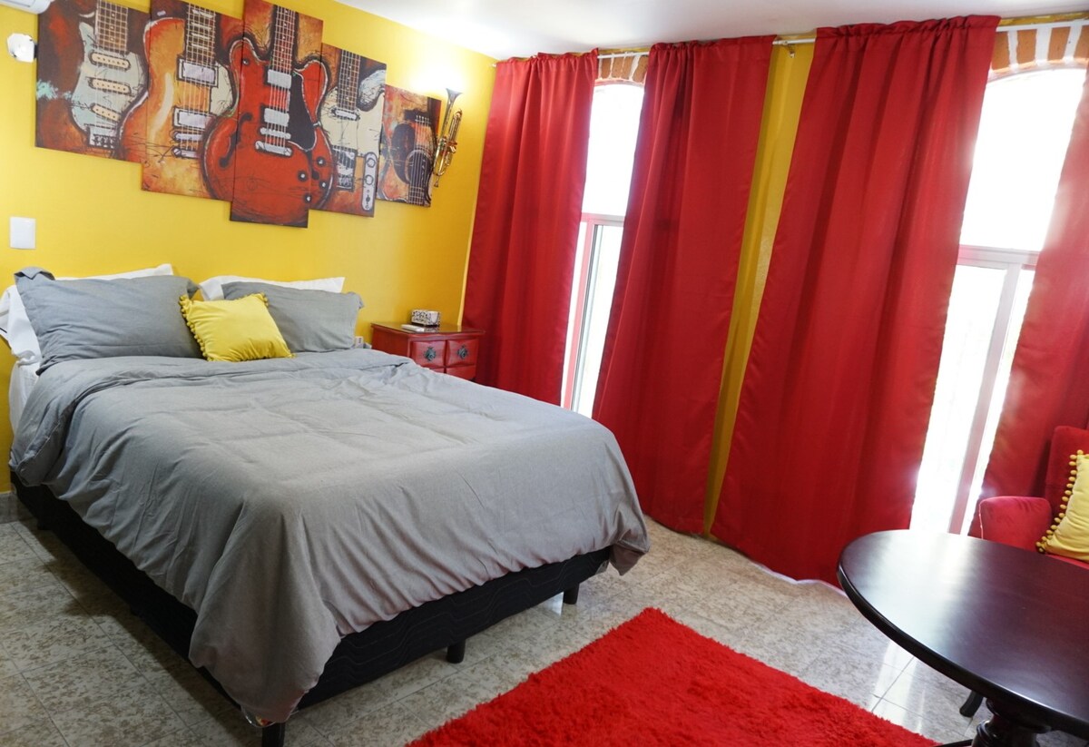 A vibrant room features a spacious bed dressed in gray linens and accented with a yellow pillow. The walls are adorned with guitar-themed artwork, while bright red curtains frame two windows, allowing natural light to enter. A small round table and red area rug complete the space.
