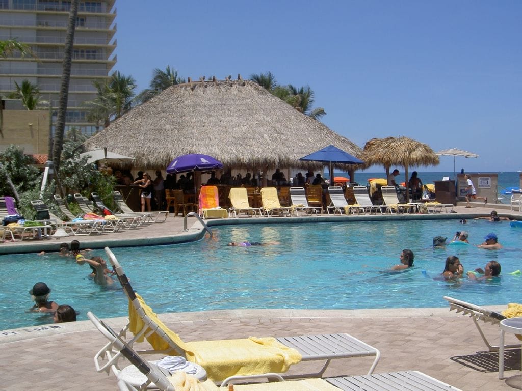 A pool area is presented with comfortable lounge chairs arranged around a clear blue pool. In the background, a thatched-roof tiki bar is visible, providing shade. Lush palm trees and the beach can be seen beyond the pool, creating a relaxed atmosphere.