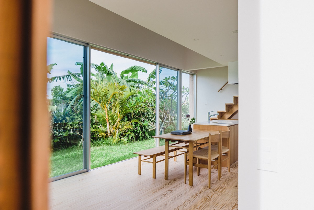 A light-filled dining area features large glass doors that open to a lush garden, allowing for a connection with nature. A wooden table and benches provide space for meals and gatherings, complemented by a staircase leading to the upper level.