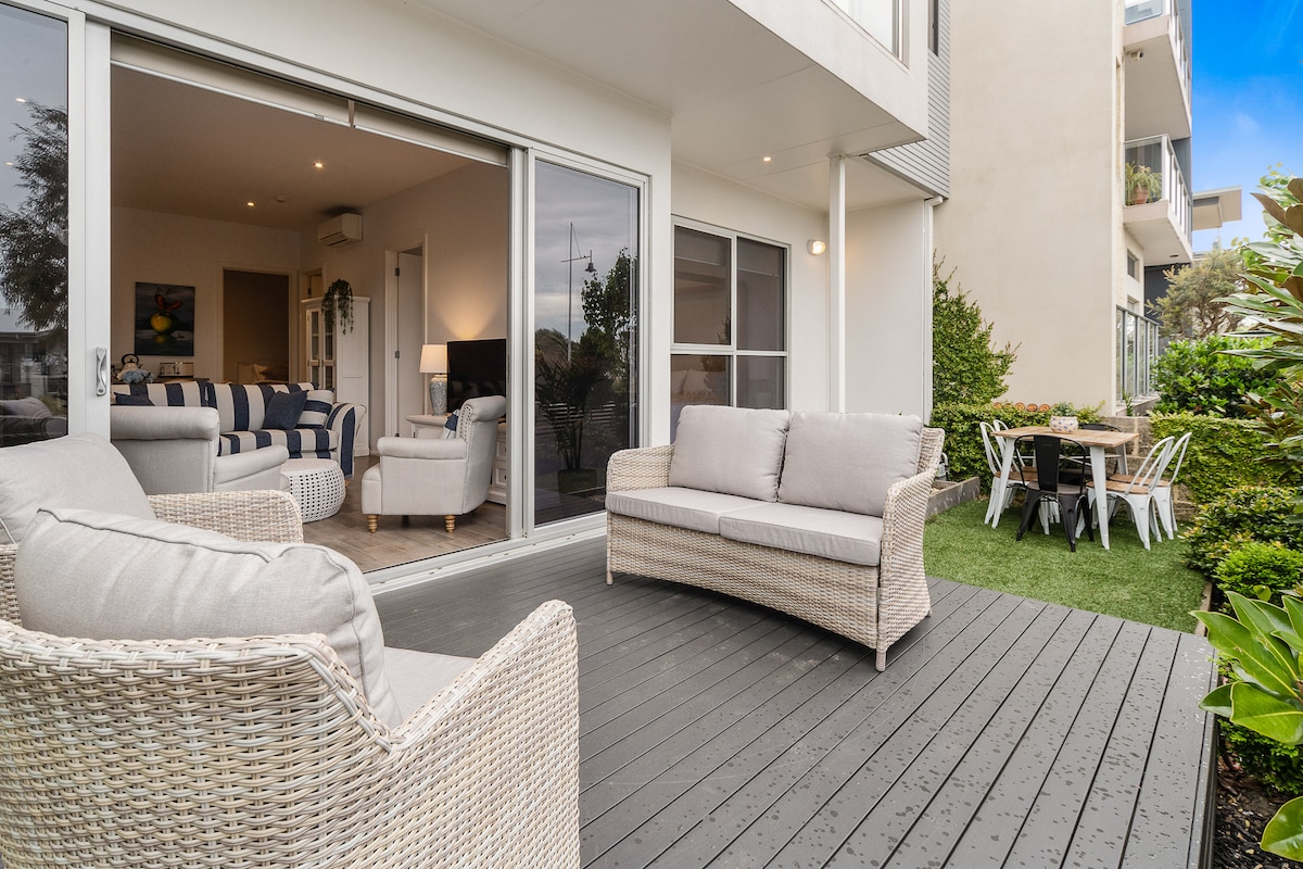 An outdoor deck area features a light-colored woven sofa and a dining table surrounded by chairs. Lush greenery lines the space, and large sliding glass doors provide a view of the open-plan living area within the apartment.