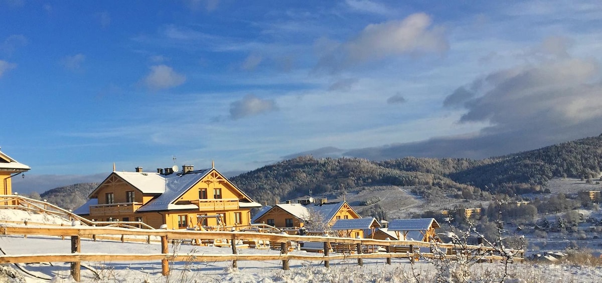 A scenic view of charming yellow cottages blanketed in snow, surrounded by a white landscape. Lush green hills in the background showcase a serene winter setting under a bright blue sky with scattered clouds.