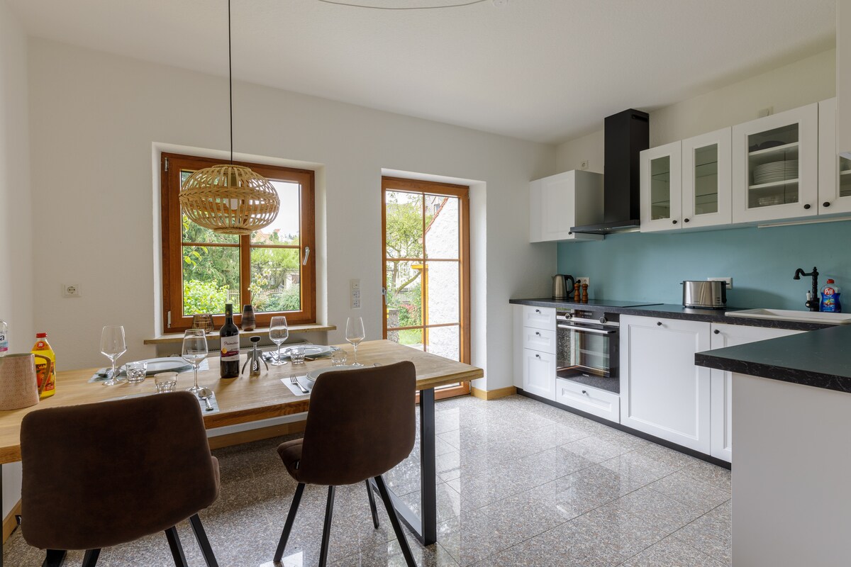 A dining area featuring a wooden table set for four, complemented by two dark chairs. The kitchen is equipped with modern appliances and has a large window allowing natural light to fill the room, offering a view of the garden.