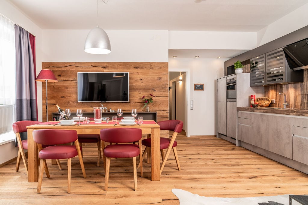A modern dining area features a wooden table surrounded by six red chairs. A large television is mounted on a wood-paneled wall, with a sleek kitchen visible in the background, showing stylish cabinetry and appliances. Natural light enters through the window dressed in curtains.