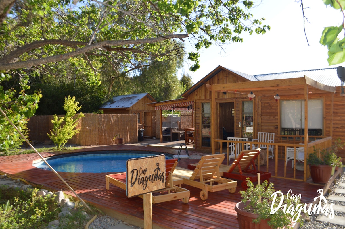 A wooden cabin is surrounded by lush greenery, featuring a private pool at its center. Sun loungers line the poolside deck, and an outdoor dining area is visible on a covered porch. A sign displayed in front of the cabin identifies the property.