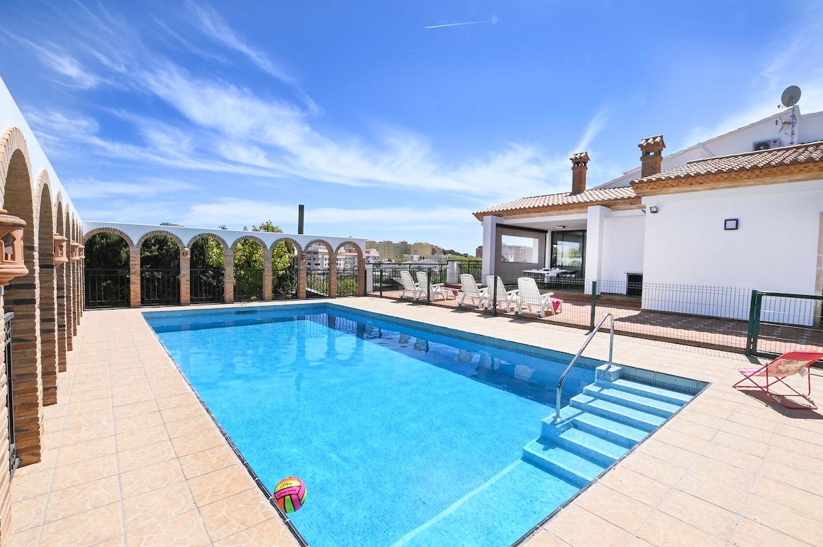 An outdoor pool area features a spacious swimming pool surrounded by sunlight. Comfortable lounge chairs are lined along one side, with shaded areas visible near the structure. A clean, tiled deck complements the serene setting, offering a perfect space for relaxation and enjoyment.