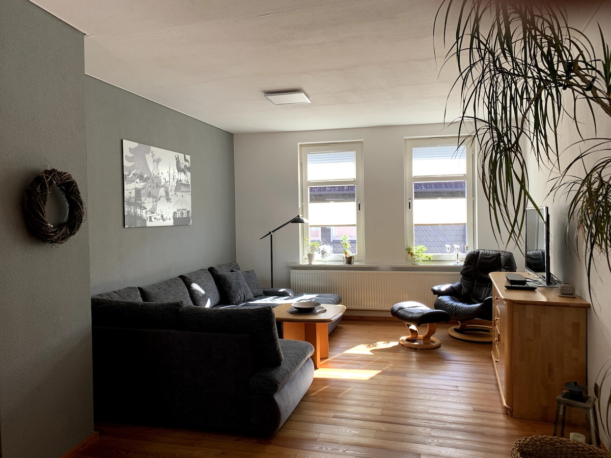 A spacious living room features a large gray sofa and a comfortable armchair positioned near a window. Light streams in from the windows, illuminating the wooden floor. A wall-mounted flat-screen TV is visible, alongside a wooden console and a decorative plant.