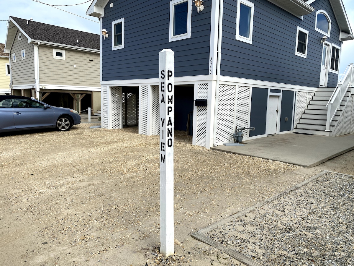 A white wooden post stands in the foreground, marked with the name 'Pompano.' The gravel driveway leads towards a two-story blue house with a garage beneath it. Additional parking spaces are visible to the side, with a second house partially seen in the background.