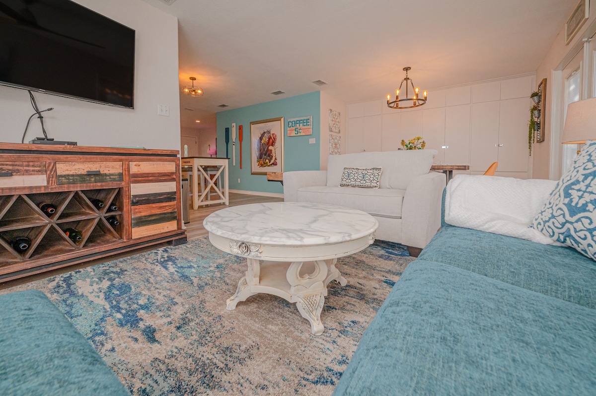A spacious living area features a light blue sectional sofa and a white marble-topped coffee table. A wooden console table with wine storage is positioned next to a wall-mounted television. Natural light fills the area, enhancing the contemporary decor.