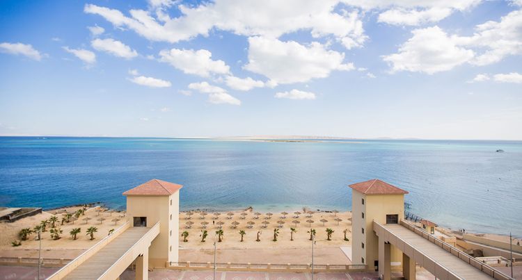 A serene coastal view stretches across the horizon, featuring gentle waves lapping against the shore. Two similarly designed towers frame the scene, with sandy beach umbrellas visible below. A clear blue sky complements the tranquil sea, creating a calming atmosphere.