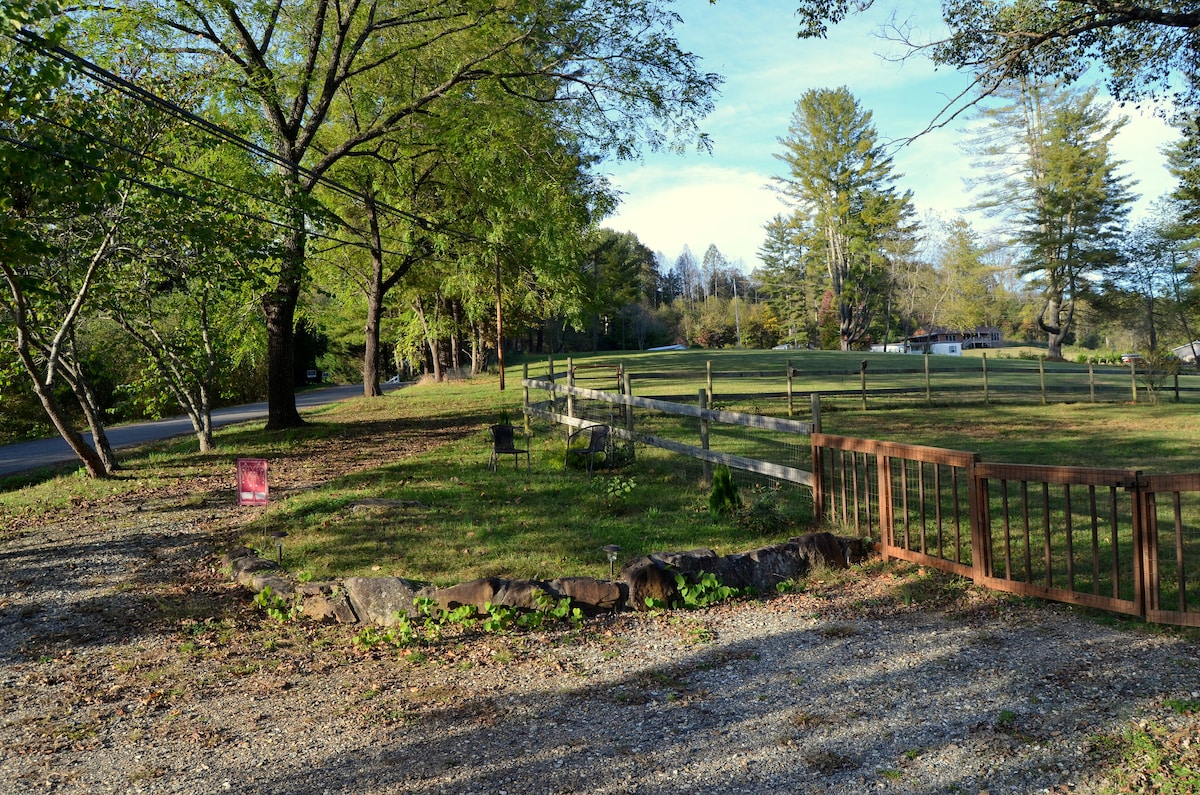An open green space is framed by wooden fencing, with the road visible in the background. Trees provide shade, and a few chairs are located nearby. A small wooden gate leads into the grassy area, creating a welcoming outdoor space for relaxation.