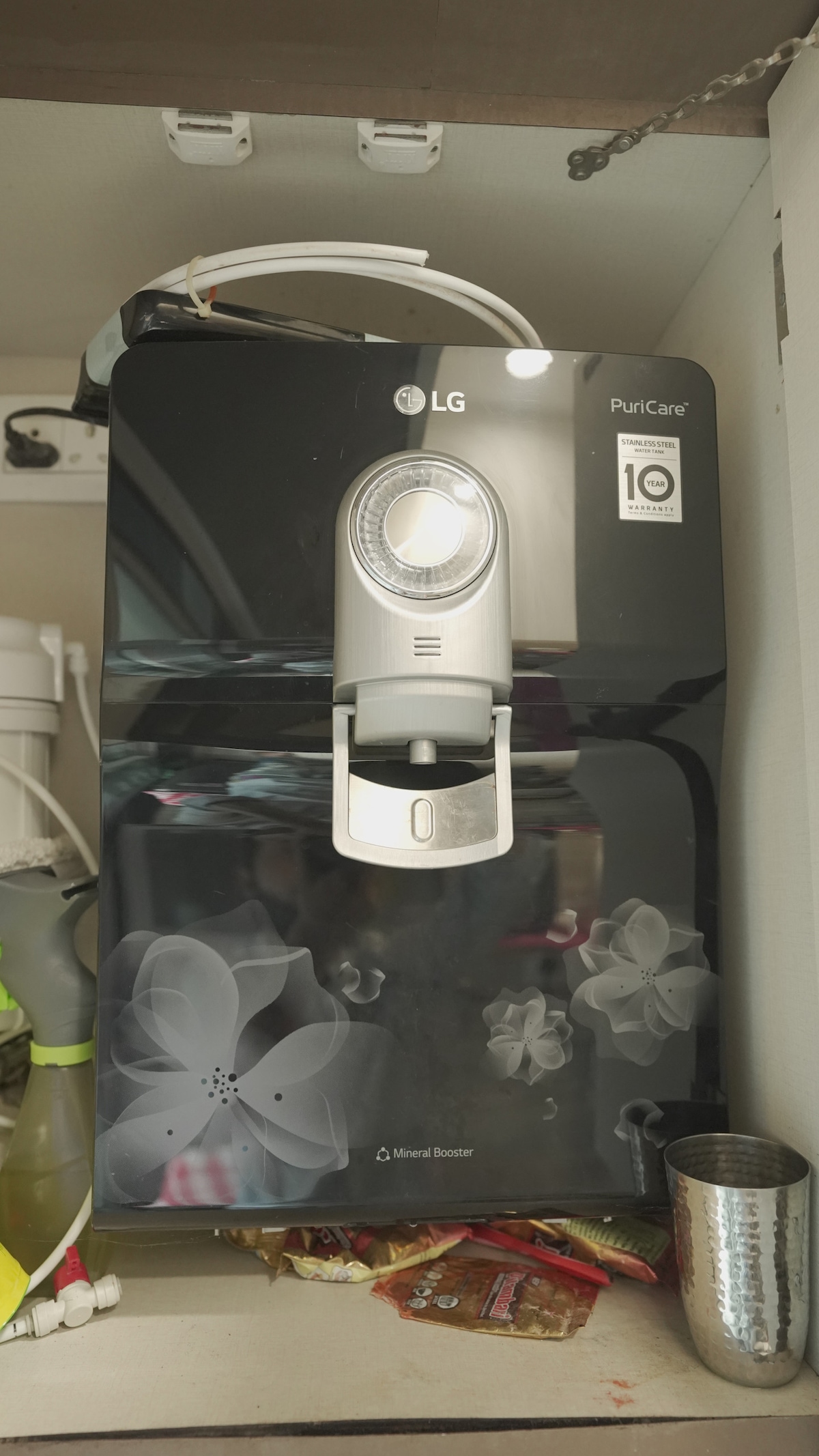 A sleek water purifier is installed on the kitchen cabinet, featuring a minimalist design with a central faucet. The dark surface is accented by floral patterns, and a clear light indicator enhances its modern aesthetic.