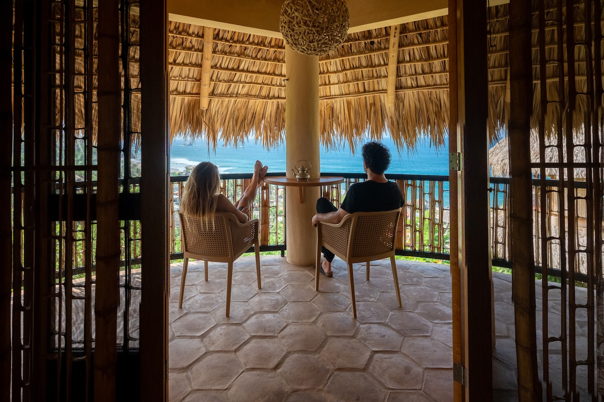 An outdoor balcony features two chairs with a natural straw roof overhead. The ocean is visible in the background, showcasing a serene view of the water and coastline. Greenery is seen below, enhancing the tranquil setting.