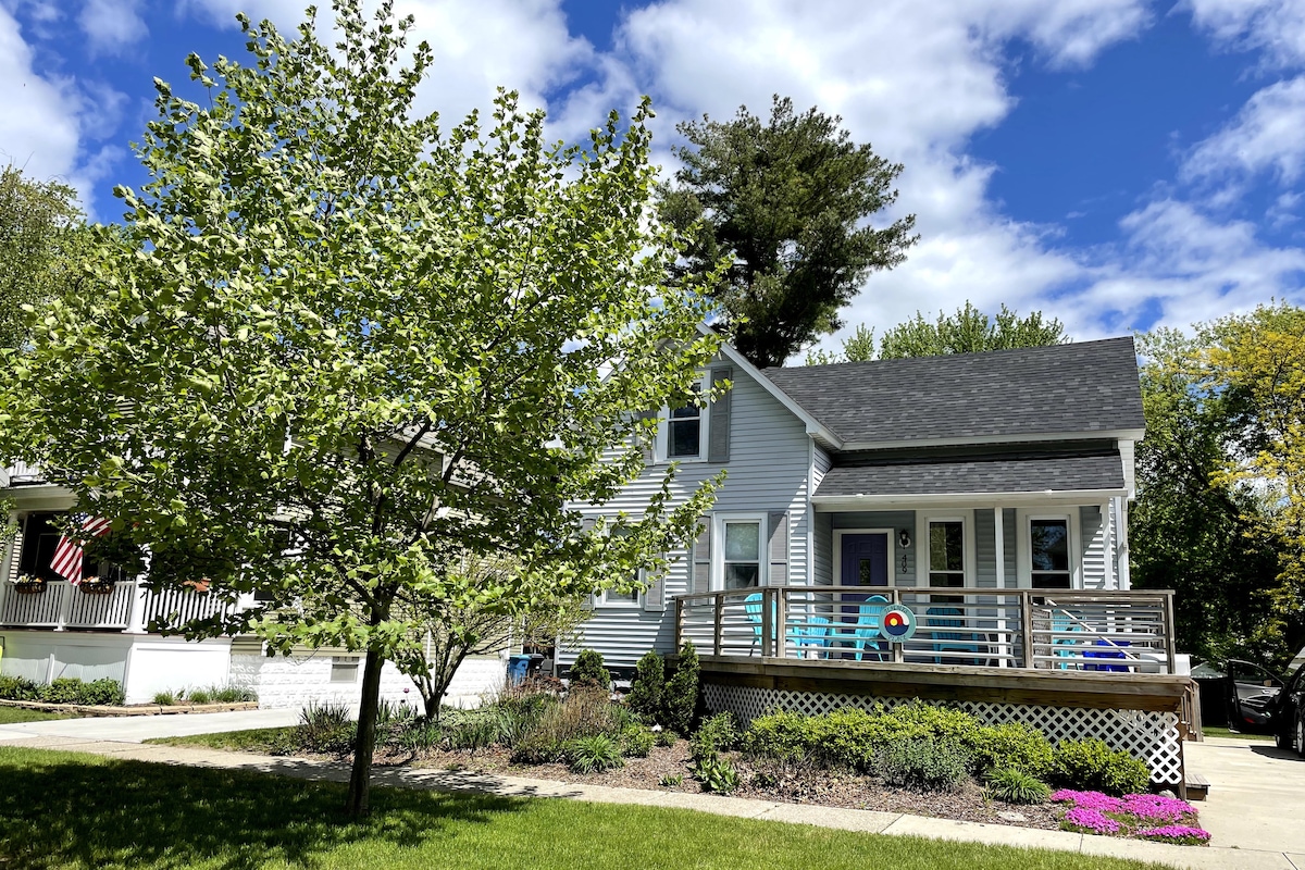 A charming two-story house is featured, surrounded by greenery and colorful landscaping. A welcoming front deck is visible, furnished with several chairs. The sky is bright with clouds, enhancing the appeal of the well-maintained exterior.
