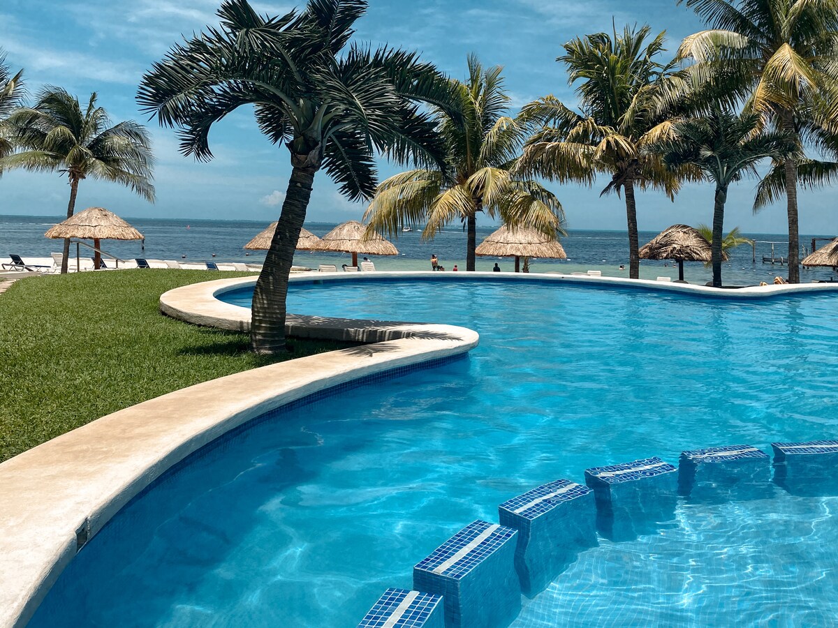 A clear blue swimming pool is surrounded by palm trees, offering a view of the ocean in the background. Beach palapas and lounge chairs are visible along the shoreline, inviting relaxation in a tropical setting.