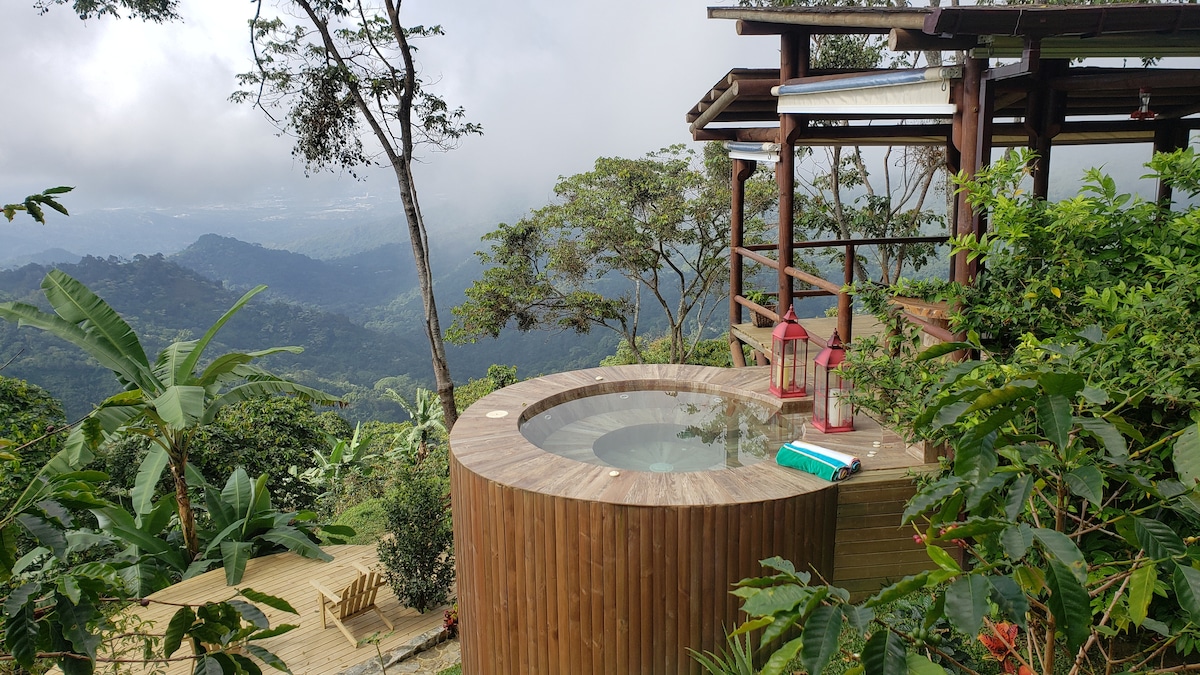 A circular hot tub is surrounded by a wooden deck and lush greenery, offering panoramic views of the mountainous landscape. Lanterns are positioned nearby, adding a touch of charm. The misty scenery creates a tranquil atmosphere, ideal for relaxation amidst nature.