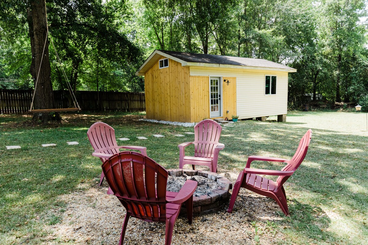 A fire pit surrounded by four maroon chairs is positioned in a spacious, grassy outdoor area. In the background, a small yellow and white building features large windows and a welcoming entrance. Tall trees provide shade, enhancing the peaceful setting.