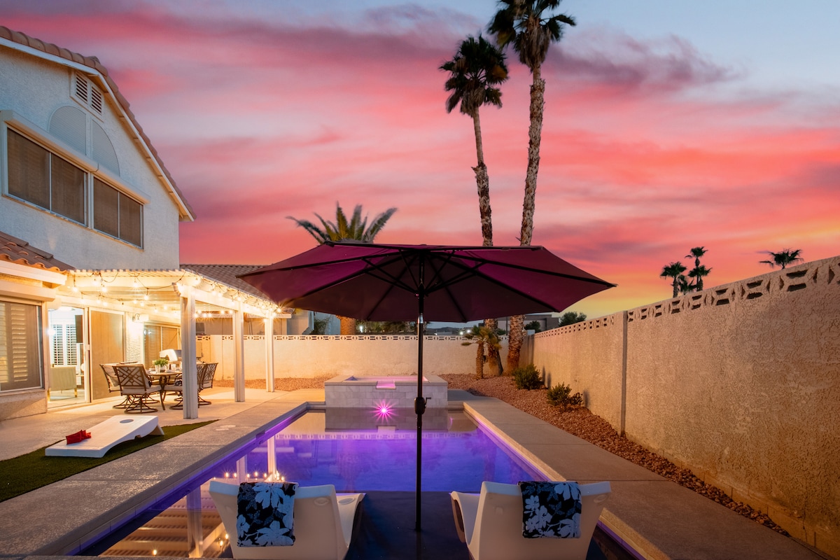 A modern backyard is featured, with a swimming pool under a vibrant sunset sky. Two lounge chairs, shaded by a large umbrella, are positioned near the pool. Palm trees frame the scene, enhancing the outdoor space designed for relaxation.
