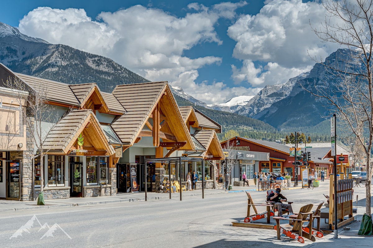 A charming streetscape showcases a row of modern buildings with wooden accents, set against a backdrop of mountainous scenery. Shops line the street, and a few people are seen enjoying the outdoors, capturing the vibrant community atmosphere.