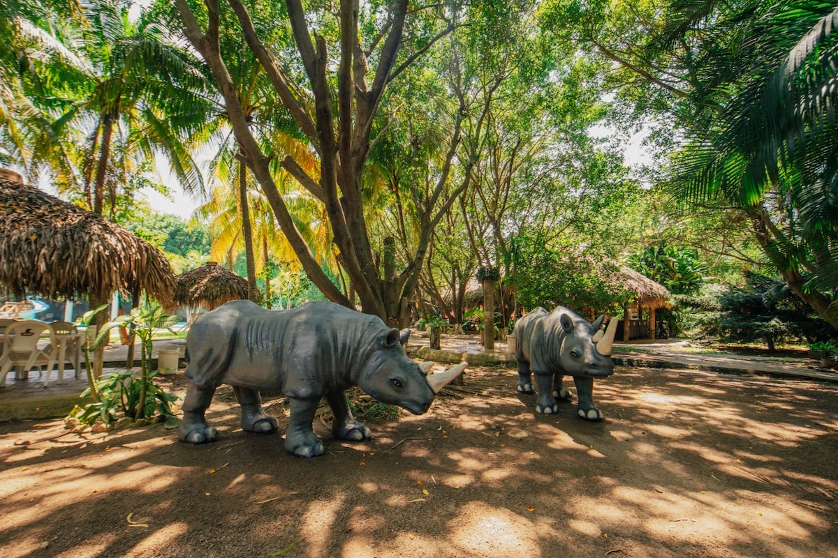 Lifelike sculptures of rhinoceroses are positioned amidst lush greenery and dappled sunlight, surrounded by tall trees and tropical plants. Thatched-roof structures are visible in the background, contributing to the natural ambiance of the area.