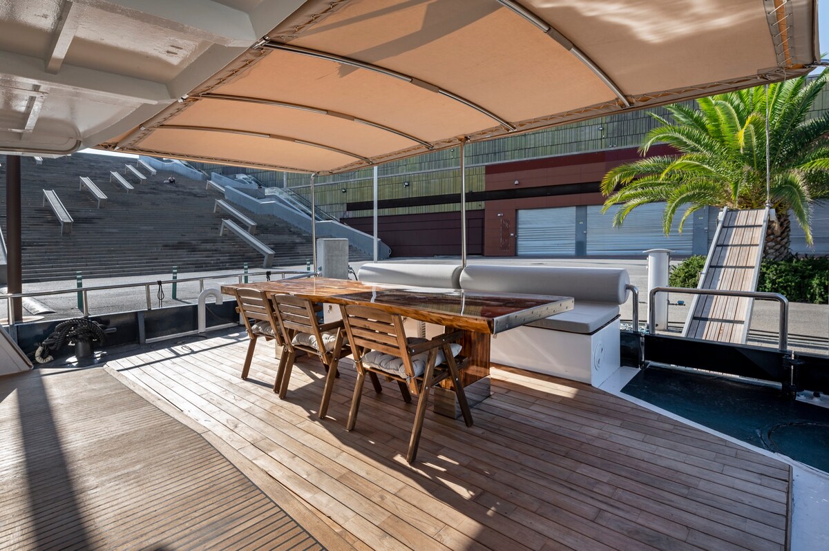 A spacious outdoor dining area features a wooden table surrounded by several chairs, set beneath a canopy for shade. The flooring is wooden, and palm trees and a ramp are visible in the background, creating a relaxed atmosphere for meals.