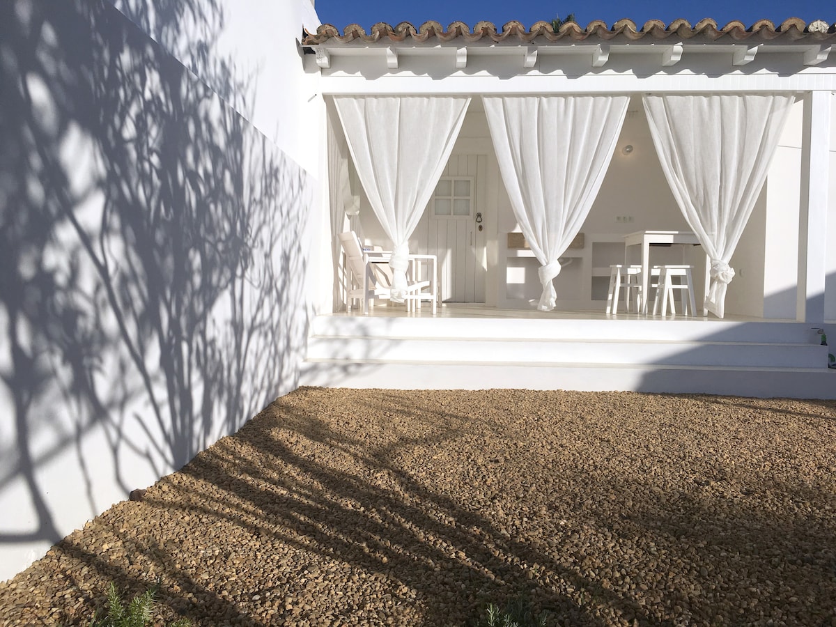 A sunlit porch is depicted, featuring flowing white curtains that softly frame light wooden furniture. A gravel area in the foreground casts intricate shadows, while the direct entrance to the room is visible in the background, enhancing the tranquil atmosphere.