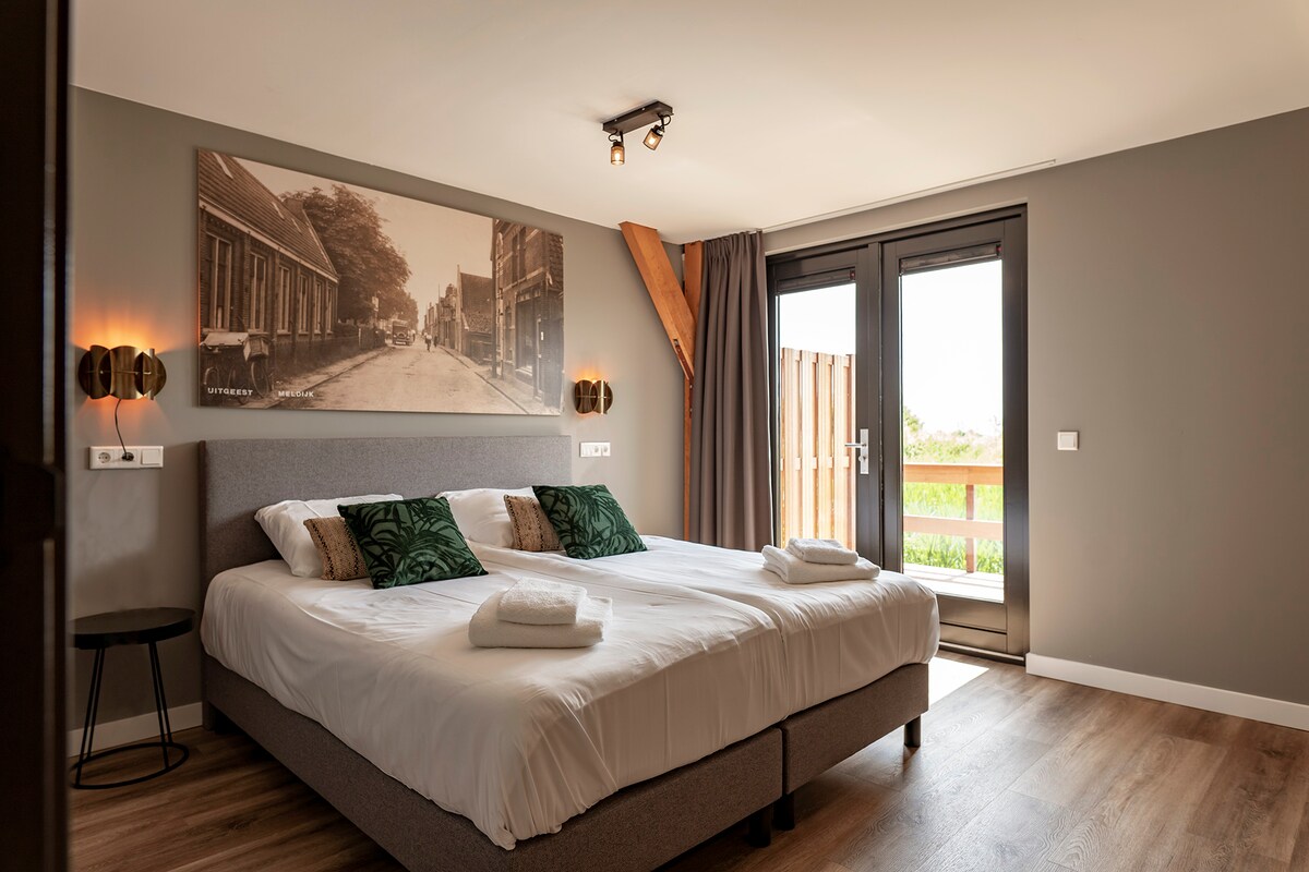 A spacious bedroom features two single boxspring beds adorned with white linens and decorative green pillows. Natural light floods in through large glass doors that lead to a private terrace, offering views of the surrounding greenery. A large art piece hangs above the beds.