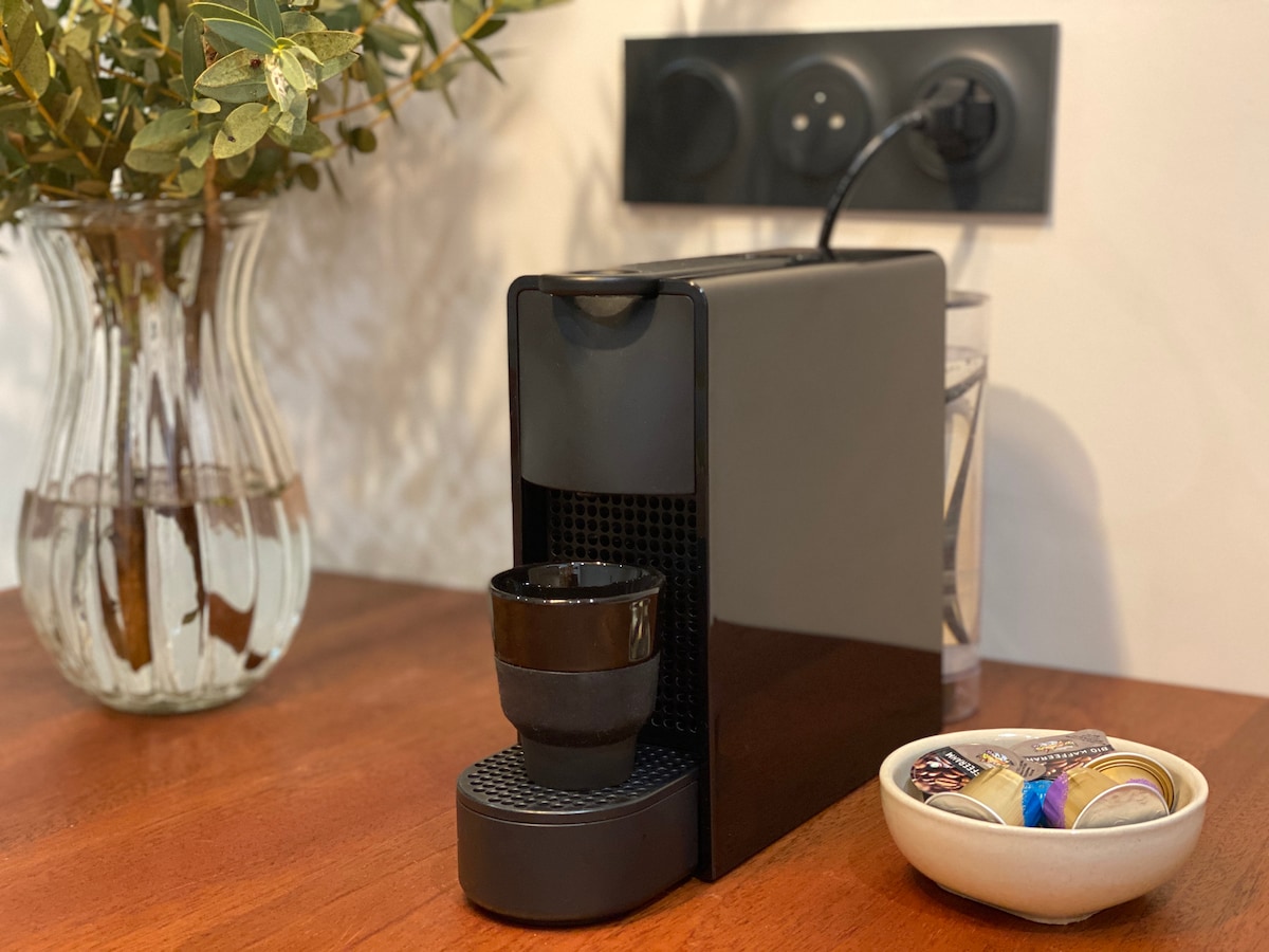 A sleek Nespresso coffee machine is positioned on a wooden countertop, next to a small dish containing coffee pods. A clear vase with greenery adds a touch of nature to the space, while the background features a wall socket that blends seamlessly with the decor.