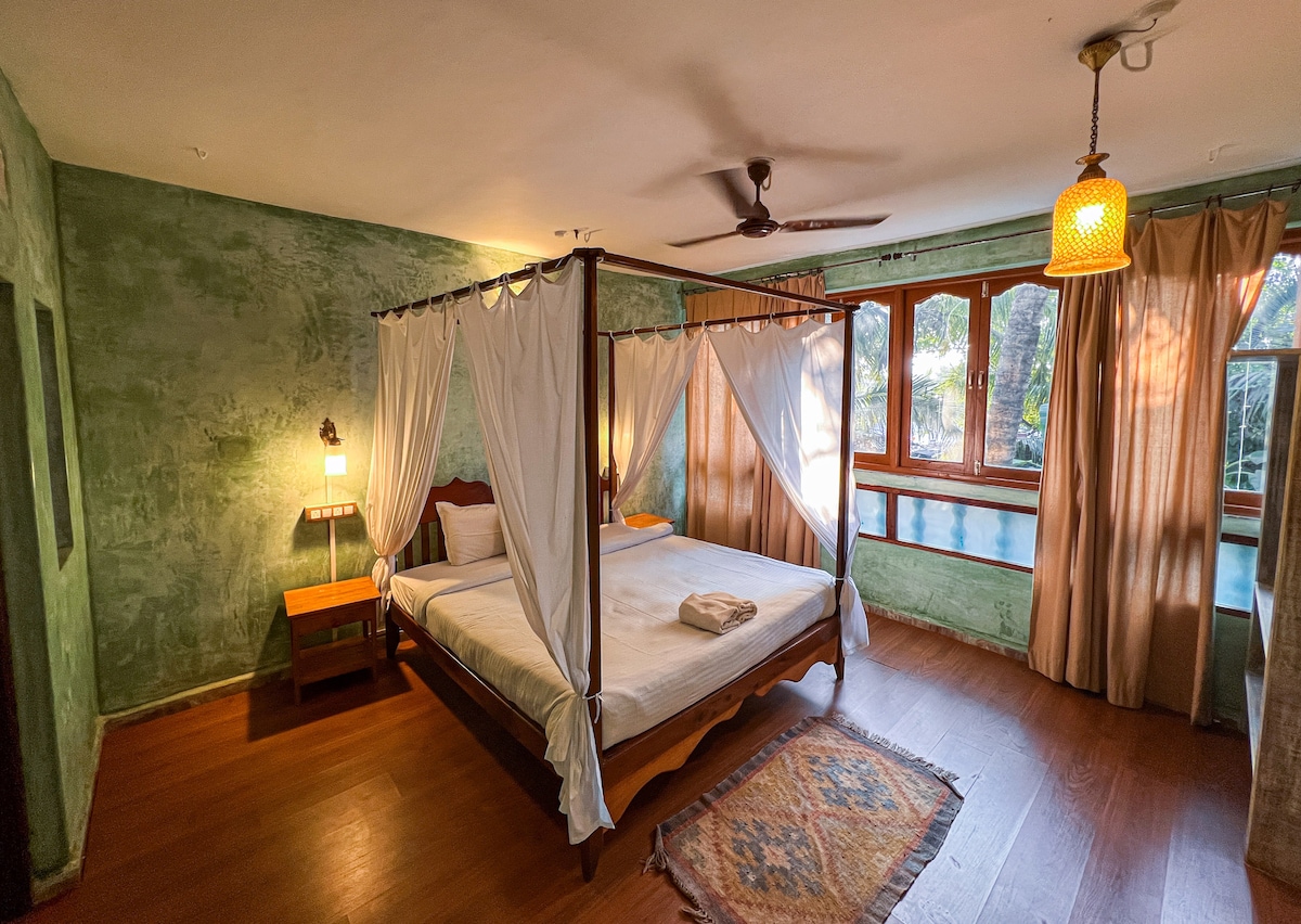 A spacious room features a canopied bed with white drapes, inviting natural light from large windows. Wooden accents and a warm color palette create a calming environment. A bedside table and a decorative rug enhance the room's charm, while greenery outside offers a soothing view.