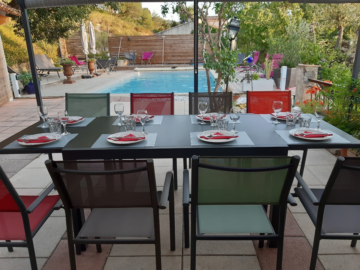 An outdoor dining table is set with plates and glasses, surrounded by colorful chairs. In the background, a tranquil pool is framed by greenery, with lounge chairs positioned for relaxation. Fresh flowers add vibrant touches to the serene atmosphere.