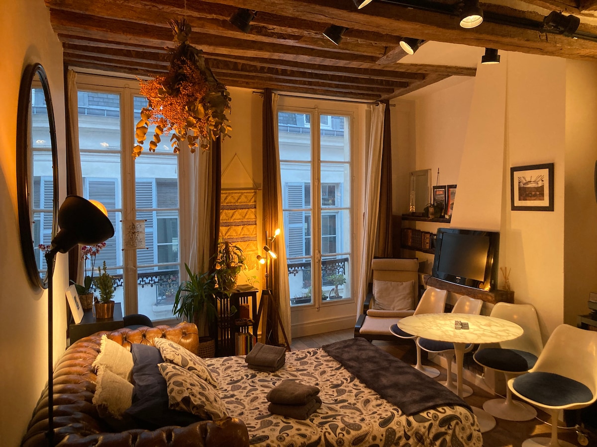 A cozy living space features a spacious bed adorned with patterned bedding. Natural light filters through large windows, highlighting wooden beams and plants. A modern television stands adjacent to a circular dining table surrounded by sleek chairs, creating an inviting atmosphere.