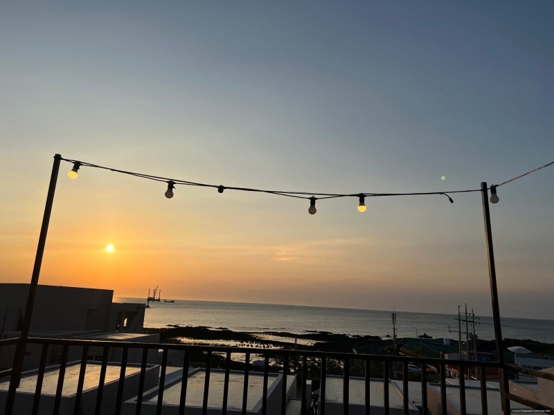 A sunset view from the terrace captures the warm hues of the sky above the ocean. Soft lighting from hanging bulbs enhances the ambiance. The horizon displays distant boats against the serene water, creating a tranquil seaside atmosphere.