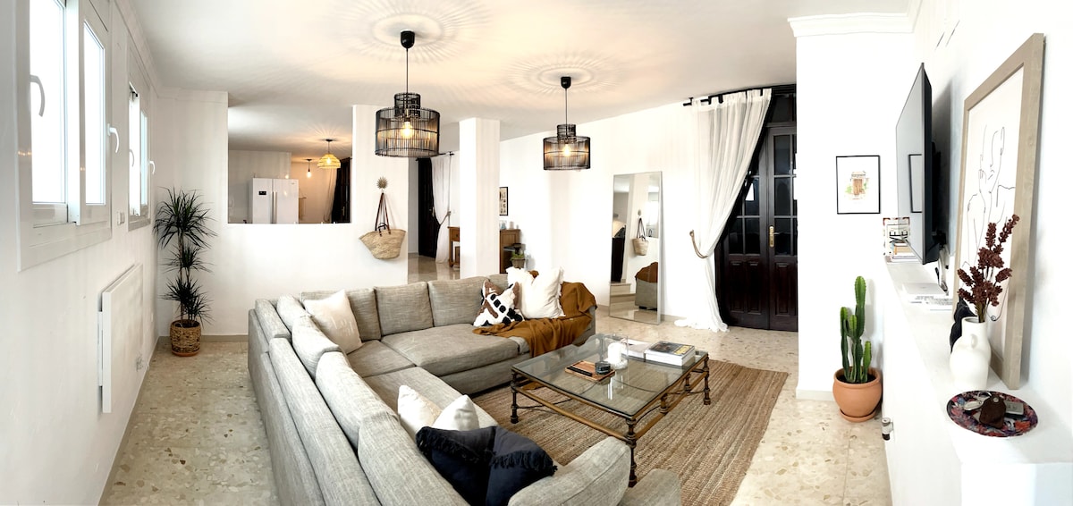 The spacious open-plan living area is filled with natural light, featuring a large sectional sofa arranged around a glass coffee table. A soft palette of white walls enhances the airy feel, while various plants bring a touch of nature to the room. Elegant lamps and decor elements add a modern touch.