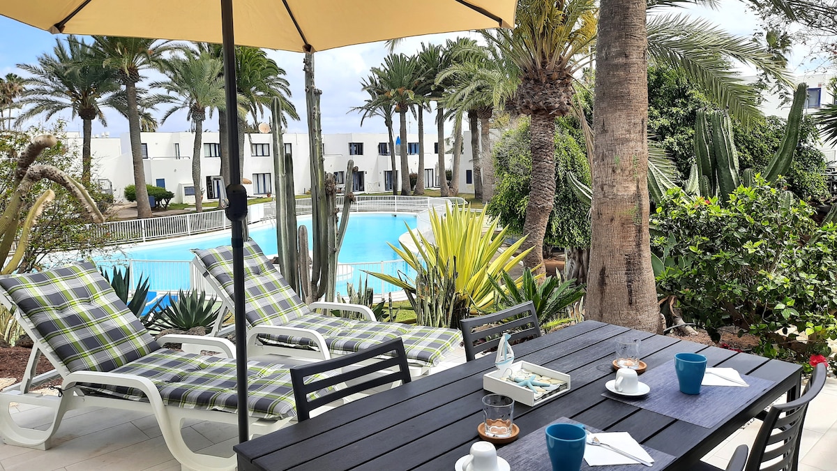 A shaded outdoor patio features a dining table set with blue cups and snacks, surrounded by lounge chairs. Lush tropical plants and palm trees frame the view of a large swimming pool, contributing to a serene outdoor environment.