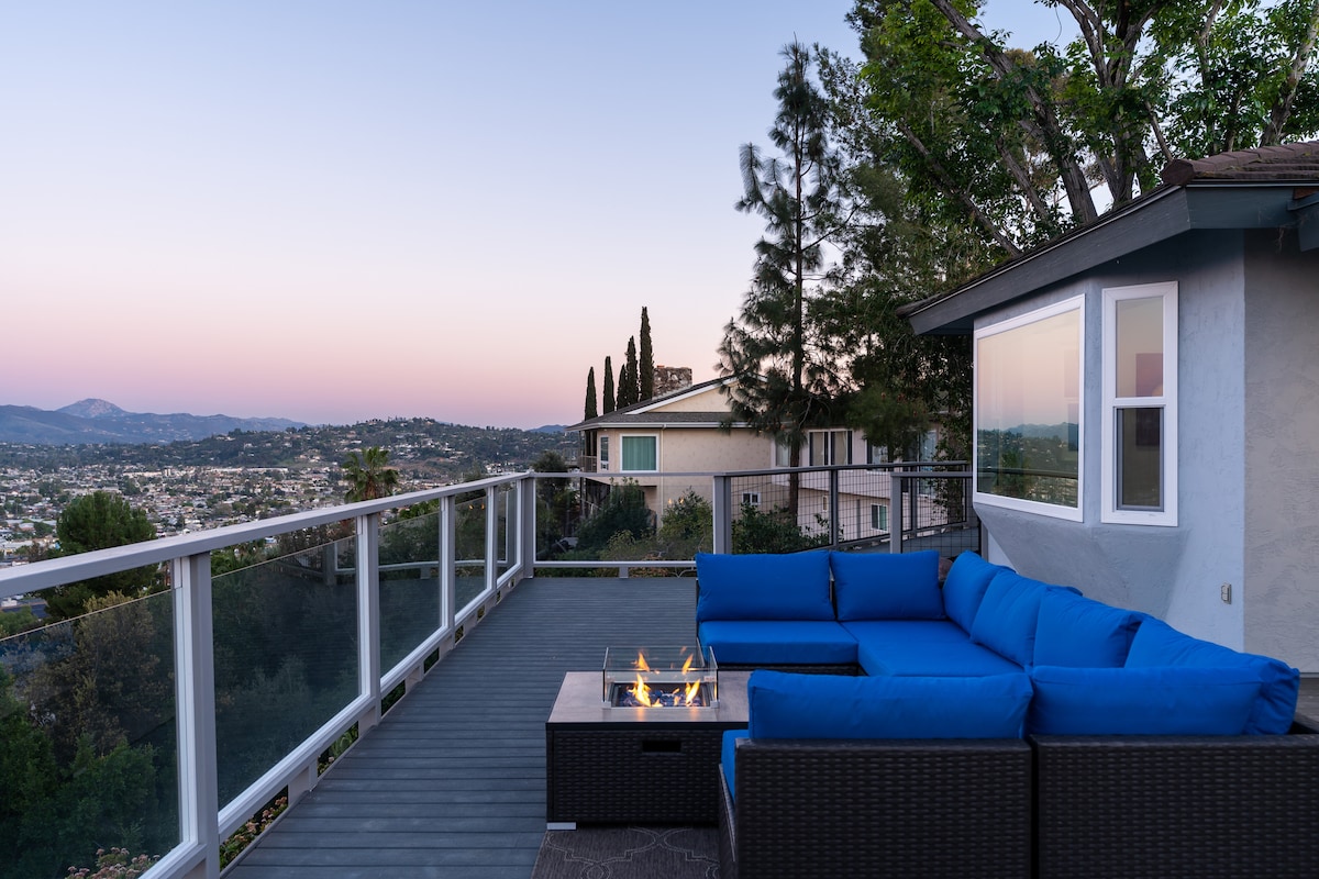 An outdoor deck features a modern seating area with a blue sectional sofa and a fire pit. Panoramic views of the surrounding landscape are visible, with trees and rooftops in the foreground. The sky transitions from soft pink to blue as dusk approaches.