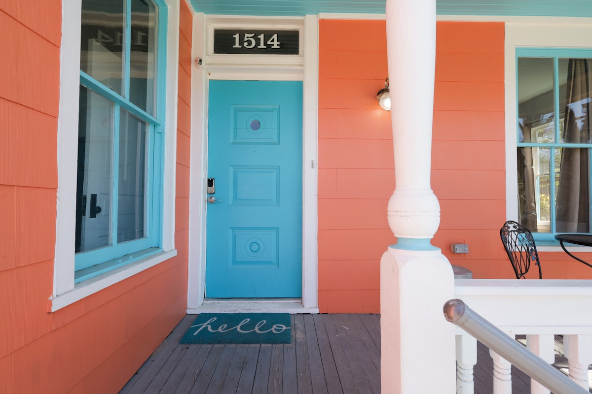 The exterior entrance features a vibrant coral façade accented by bright turquoise trim and a welcoming blue door. A small mat with the word 'hello' is placed at the entryway, while a seated area can be seen to the right, inviting guests to relax outdoors.