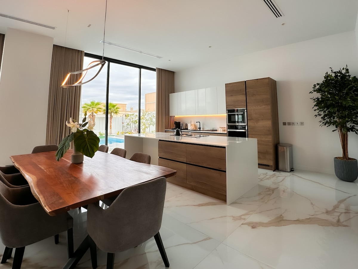 A modern kitchen and dining area are featured, with a spacious wooden dining table surrounded by upholstered chairs. The kitchen includes sleek cabinetry and stainless steel appliances. Large glass doors allow natural light and offer views of the outdoor pool area.