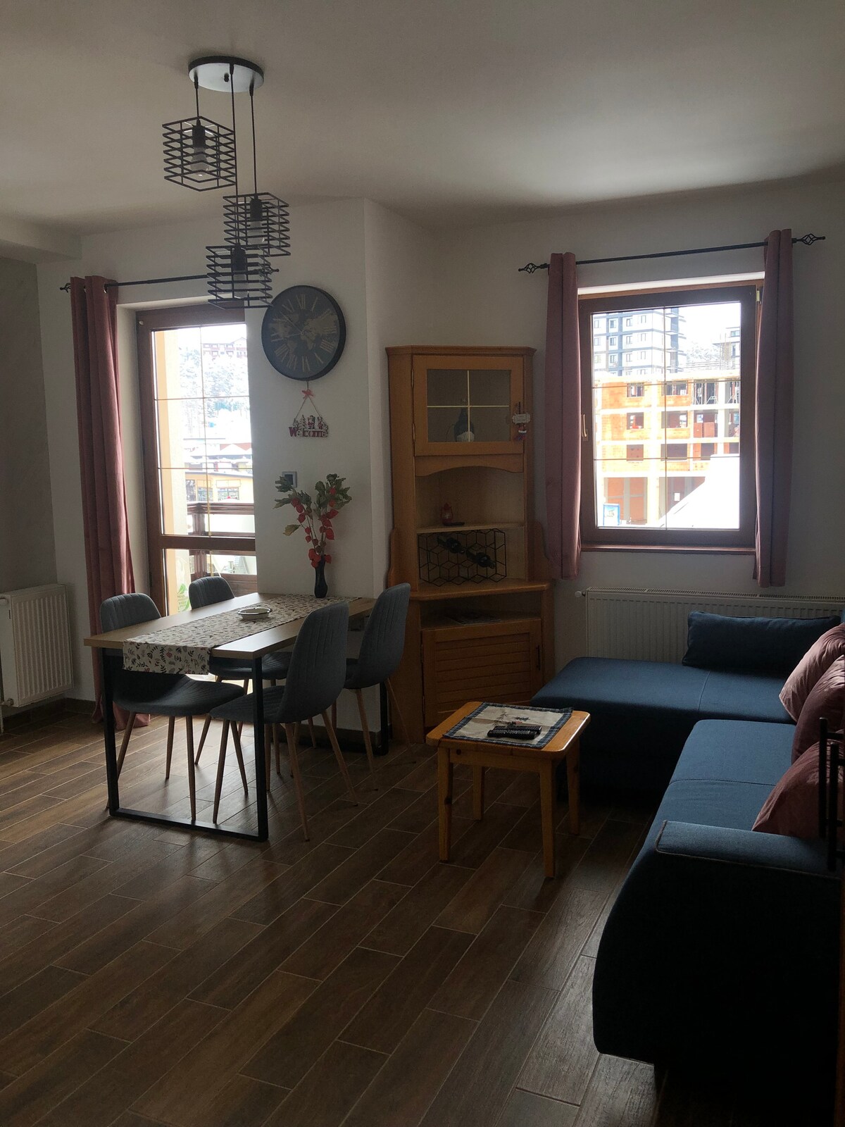 A cozy living area is presented, featuring a blue sectional sofa and a small table with four chairs. Two large windows allow natural light to fill the space, framed by light curtains. A wooden cabinet is visible beside the dining area.
