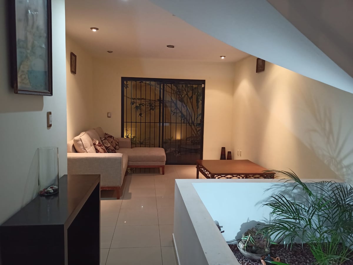 A spacious living area is illuminated by recessed lighting, featuring a light-colored sofa and a wooden coffee table. A large window with security bars offers a view of greenery outside. Decorative plants add a touch of nature to the ambiance.