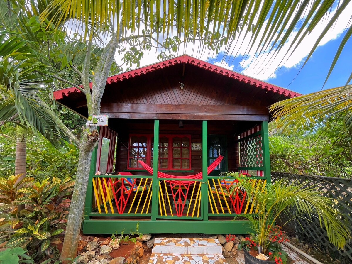 A colorful wooden cottage is surrounded by lush tropical foliage. The front porch features red and yellow chairs with hammocks, inviting relaxation. Decorative lattice accents frame the entry, and a vibrant red roof complements the charming design under a clear blue sky.